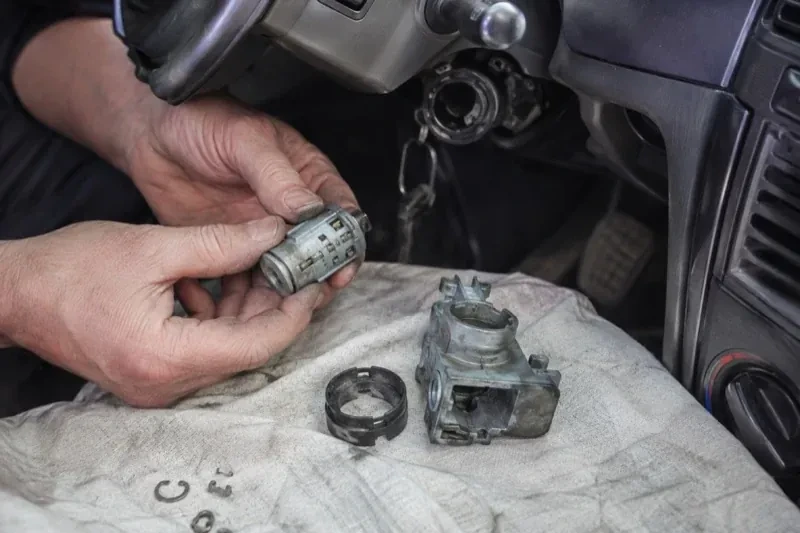 Hands holding a removed ignition cylinder beside a car steering column, with the housing and small parts on a cloth. — Ignition Lock Repair, East Gwillimbury